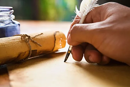 Một bàn tay cầm bút lông ngỗng chấm mực viết lên giấy da; bên cạnh là cuộn da buộc dây và lọ mực xanh.